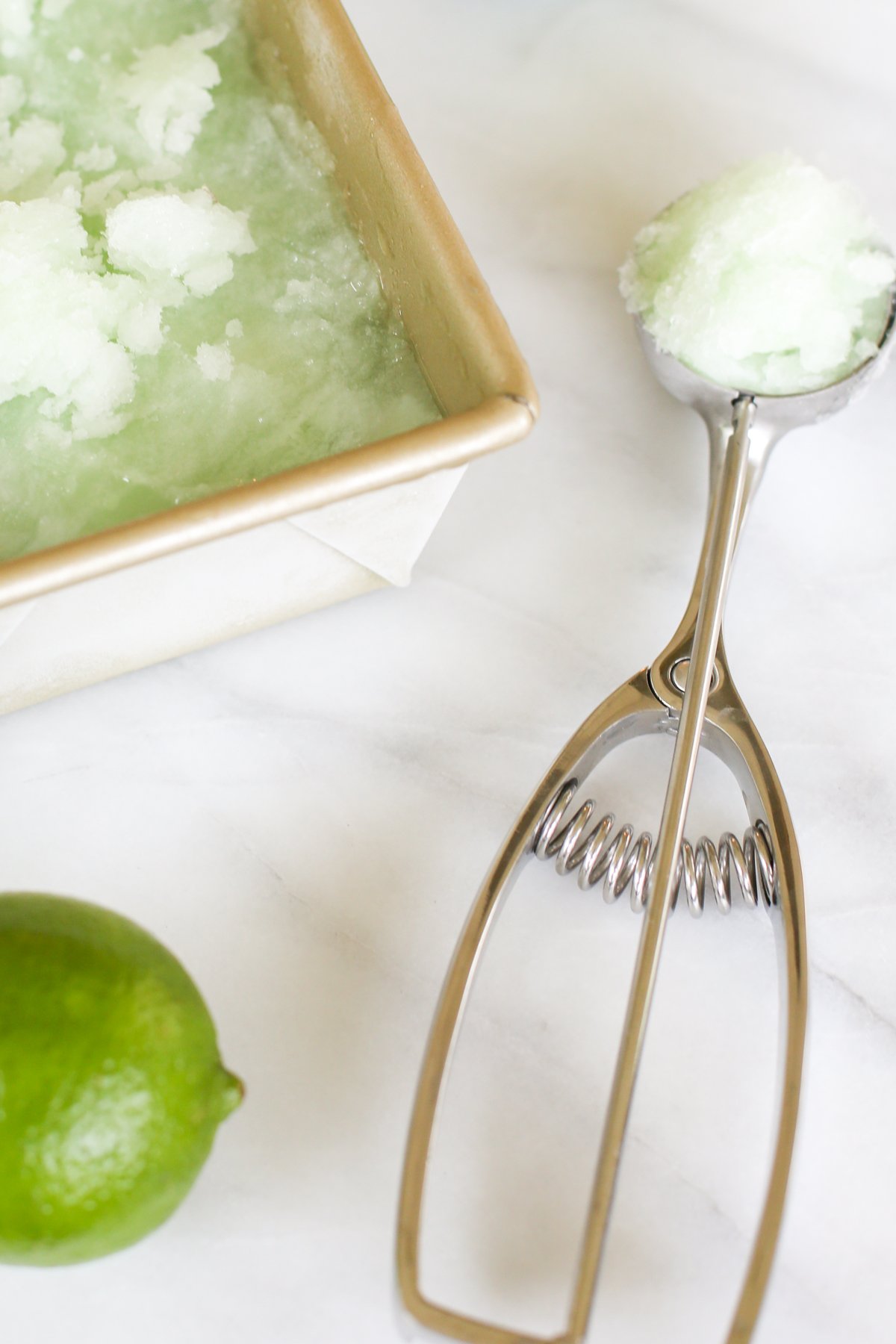 Homemade lime sorbet in a gold loaf pan. A lime and an ice cream scooper are nearby.