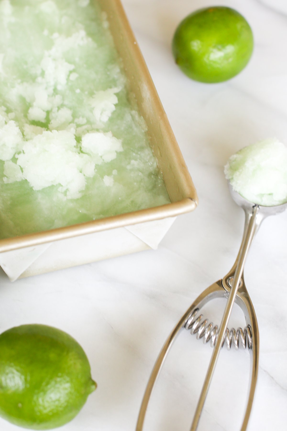 Homemade lime sorbet in a gold loaf pan. A lime and an ice cream scooper are nearby.