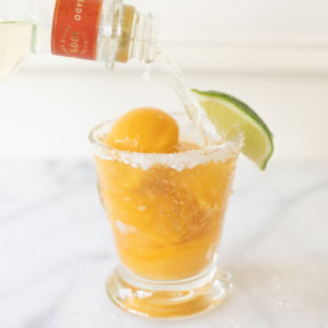 A glass of salt-rimmed mango margarita with a lime wedge on the rim is being filled with a clear liquid from a bottle, creating the perfect margarita float.