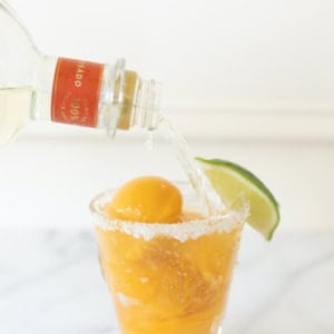 A glass with an orange drink and ice on a marble surface is garnished with a lime wedge. Clear liquid is being poured into the glass from a bottle, creating a refreshing margarita float.