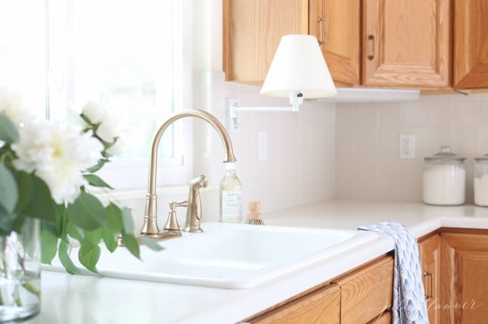 Modern kitchen with oak cabinets, champagne bronze faucet, original tile backsplash and original white porcelain sink. White flowers on counter.