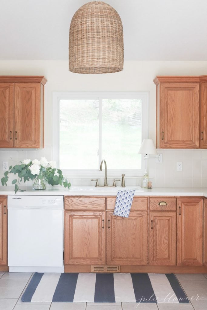 90s kitchen with oak cabinets looks modern and fresh with rattan light fixture, champagne bronze faucet, and blue and white striped rug in front of sink. 