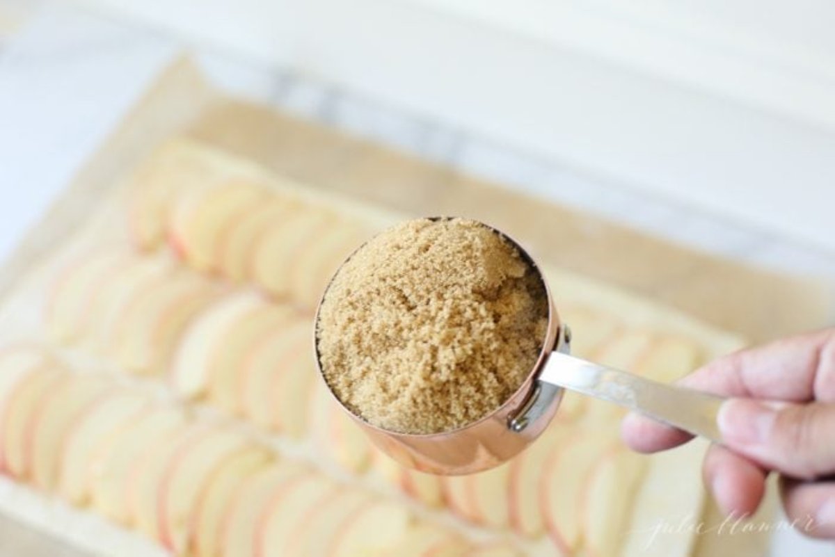 A hand holding a cup of brown sugar over a sheet of sliced apples
