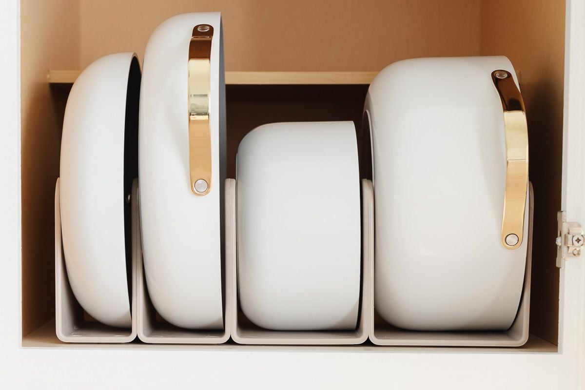 White Caraway cookware in a pots and pans organizer inside a kitchen cabinet.