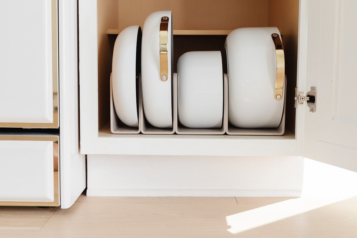 White Caraway cookware in a pots and pans organizer inside a kitchen cabinet.