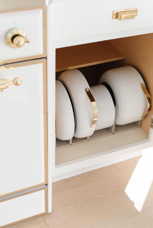 White Caraway cookware in a pots and pans organizer inside a kitchen cabinet.