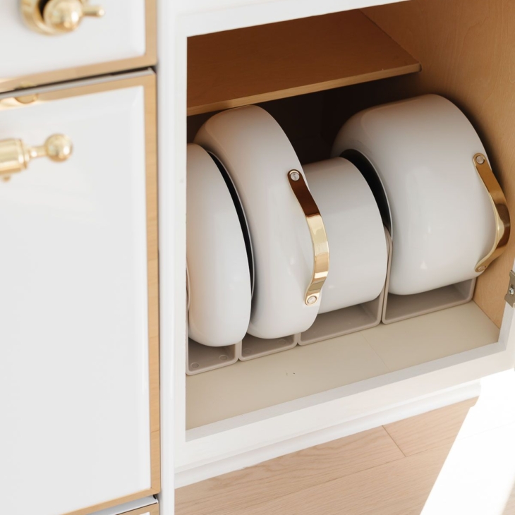 White Caraway cookware in a pots and pans organizer inside a kitchen cabinet.