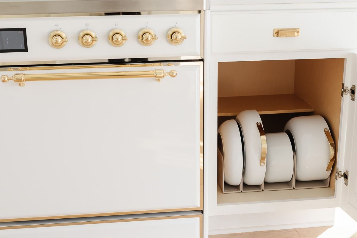 White Caraway cookware in a pots and pans organizer inside a kitchen cabinet, next to a white and gold Italian range. 