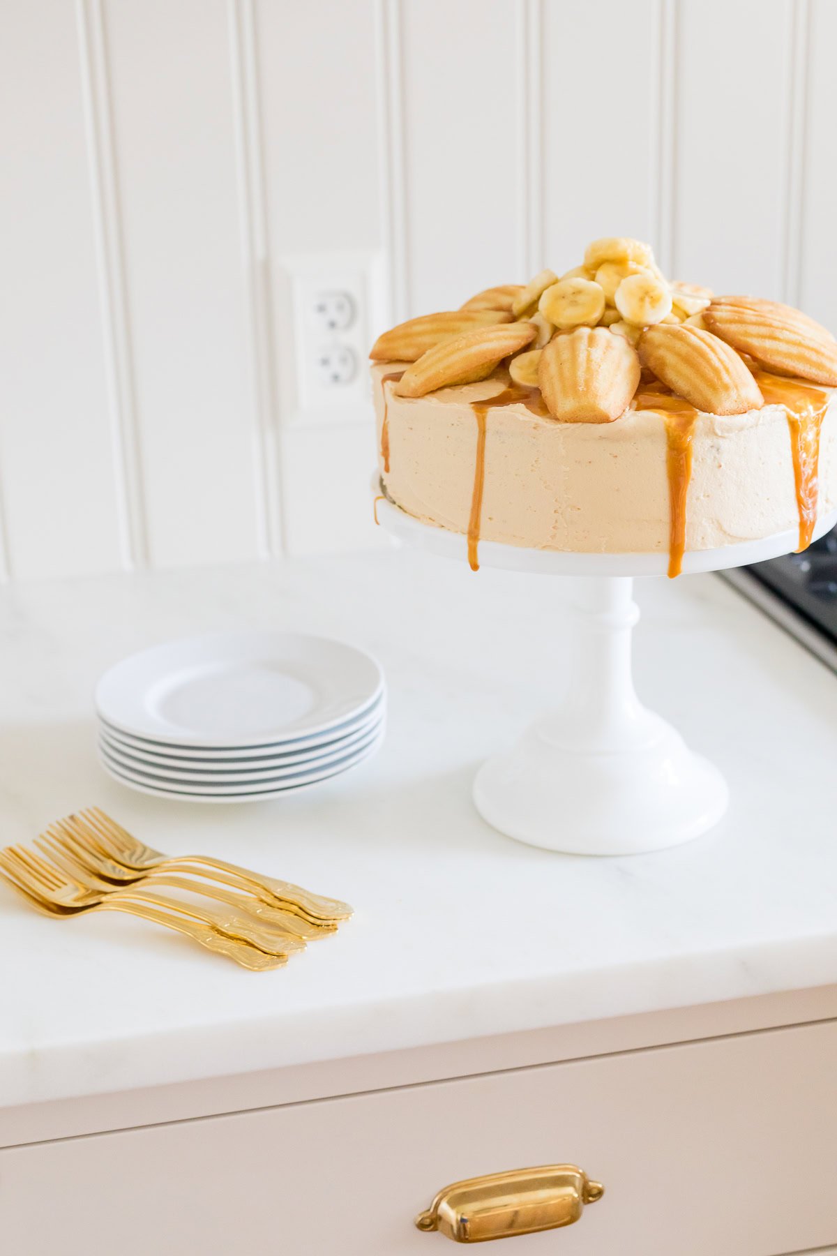 A cake with salted caramel frosting, topped with madeleine cookies and banana slices, is displayed on a white cake stand. Nearby, there are stacked plates and gold forks on a counter.