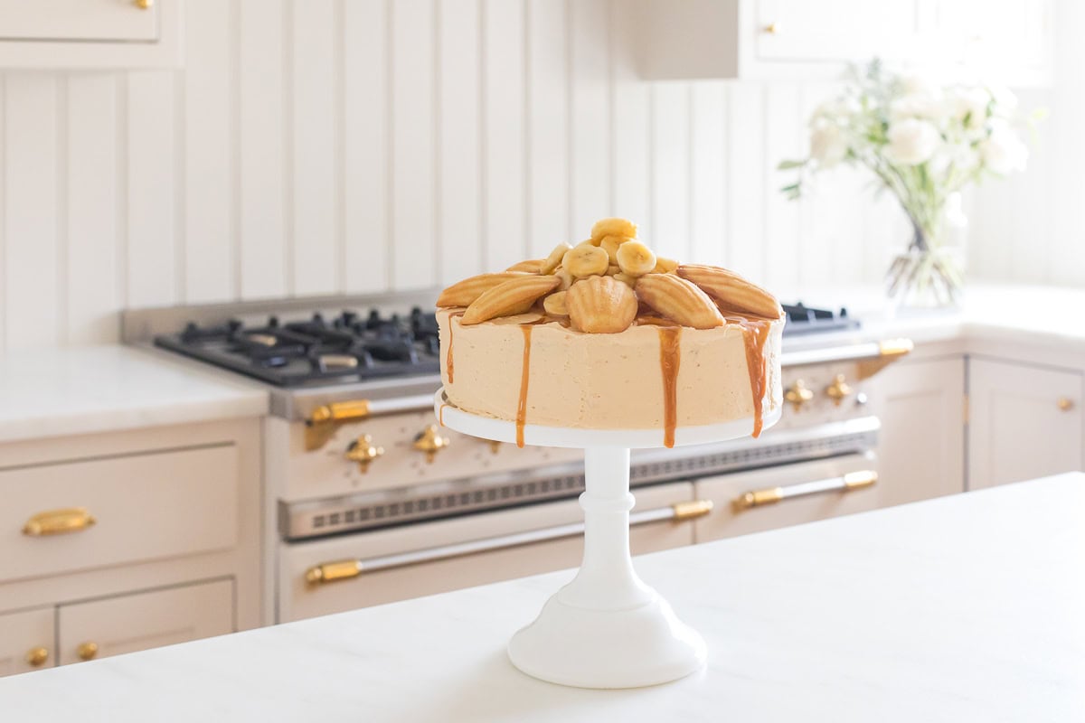 A cake on a white pedestal topped with madeleines and bananas, adorned with easy caramel frosting, sits on a kitchen counter in front of a stove, adjacent to a vase of flowers.