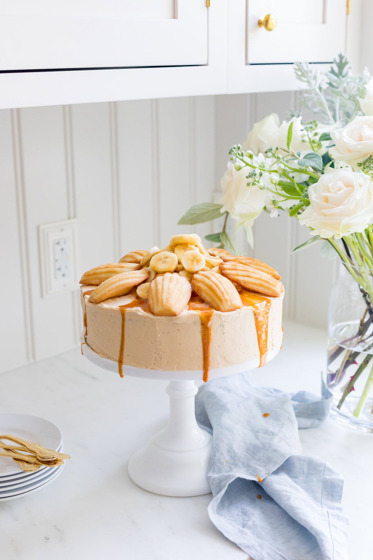 A caramel-drizzled cake topped with madeleines and banana slices, adorned with a layer of salted caramel frosting, sits on a white cake stand beside a vase of white flowers in a kitchen setting.