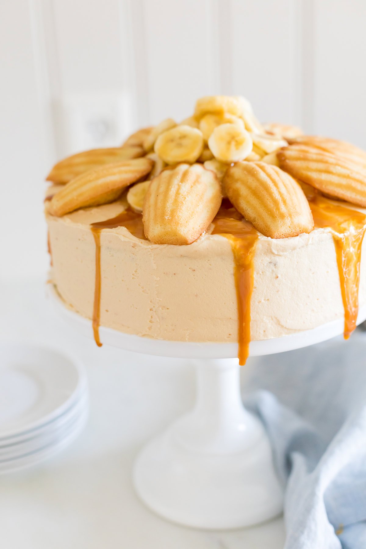 A round cake with salted caramel frosting, topped with bananas and madeleine cookies, sits on a white cake stand. Stacked plates are visible in the background.