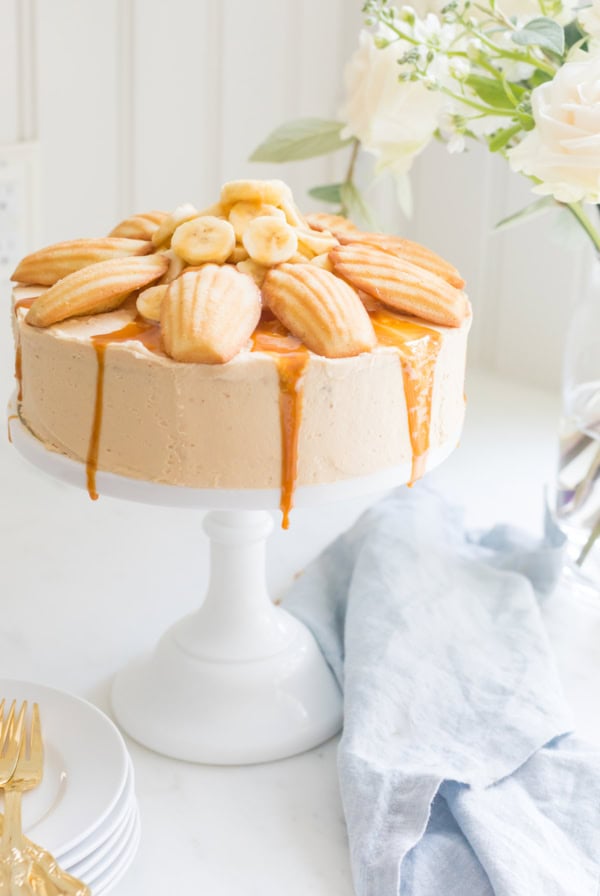 A caramel-drizzled cake topped with banana slices and madeleines sits on a white cake stand next to a stack of plates, gold forks, and a light blue cloth napkin. Flowers are in the background, complementing the homemade charm of this dessert adorned with easy caramel frosting.