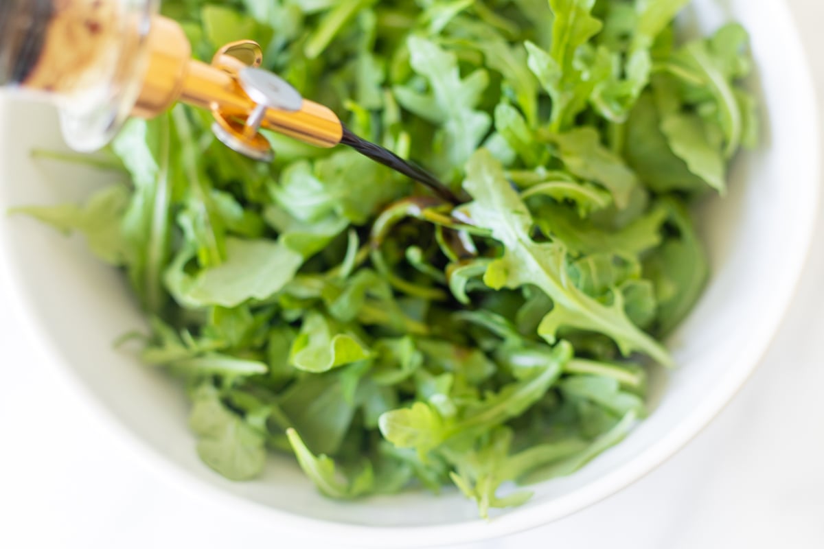 Balsamic vinaigrette in a glass bottle, pouring over an arugula salad.