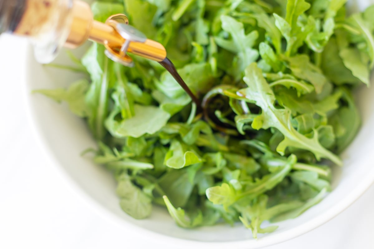 Balsamic vinaigrette in a glass bottle, pouring over an arugula salad.