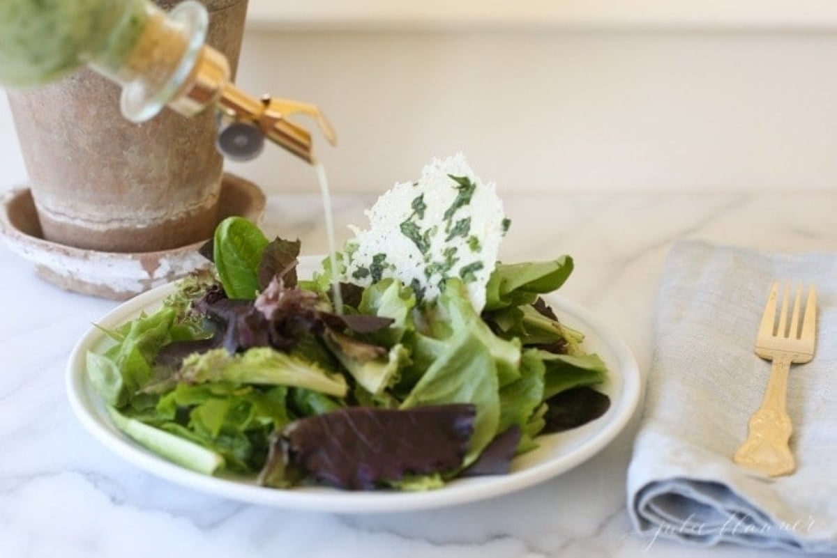 A leafy green salad on a white plate, with a glass bottle of herb vinaigrette pouring over it
