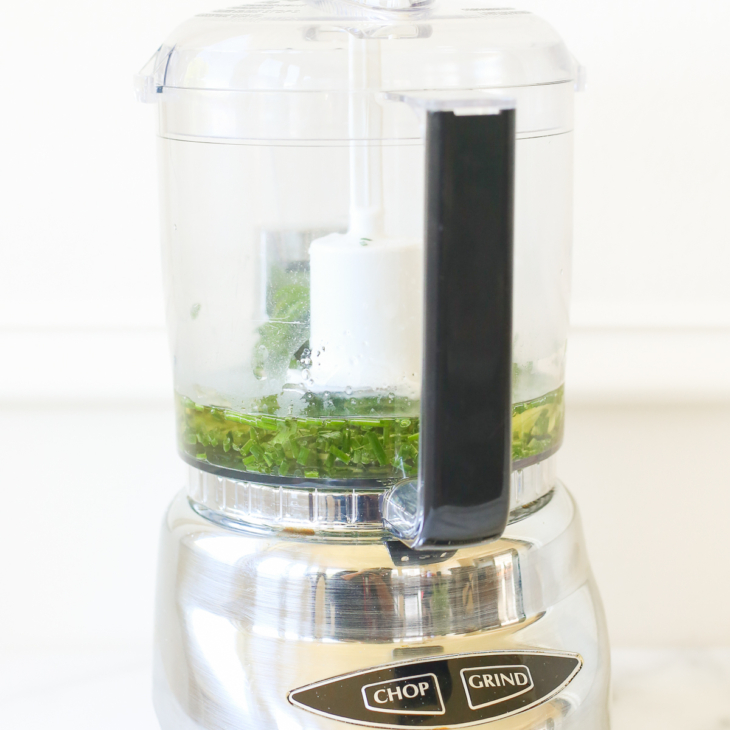 A stainless steel food processor with fresh herbs chopped in the bowl.