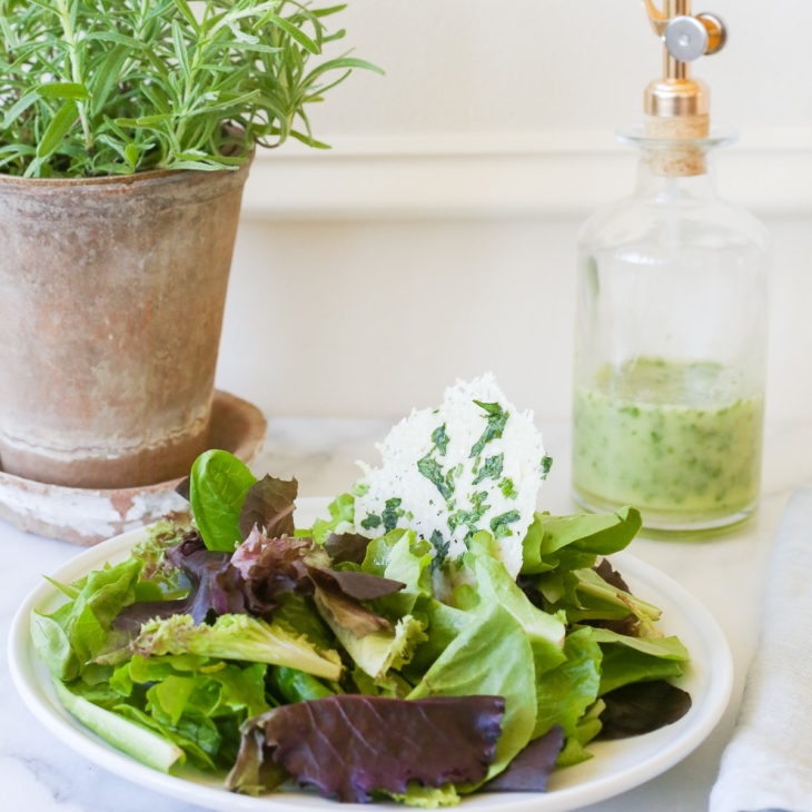 A leafy green salad on a white plate, with a glass bottle of herb vinaigrette in the background.