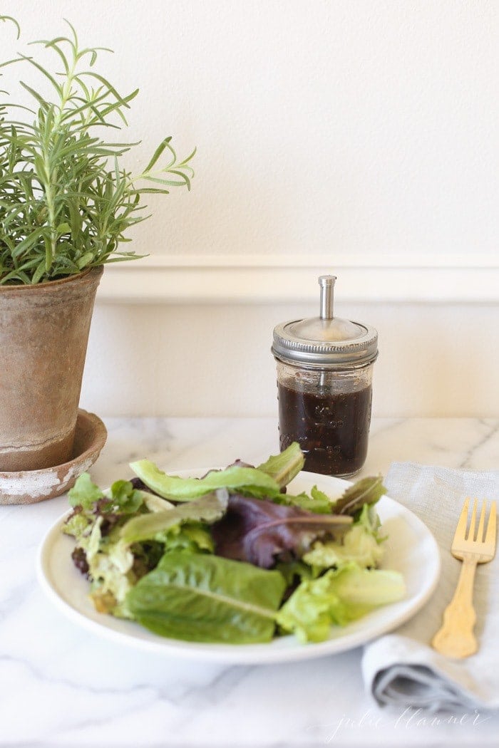 Balsamic vinaigrette in a glass mason jar with a spout for serving, placed behind a leafy green salad on a white plate.