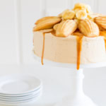A frosted cake topped with madeleines, banana slices, and a drizzle of salted caramel frosting, placed on a white cake stand, with a stack of white plates in the background.