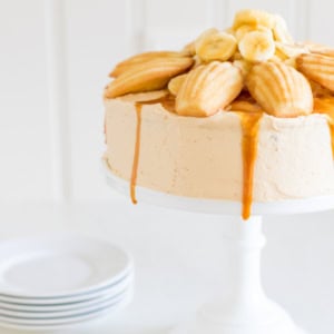 A frosted cake topped with madeleines, banana slices, and a drizzle of salted caramel frosting, placed on a white cake stand, with a stack of white plates in the background.