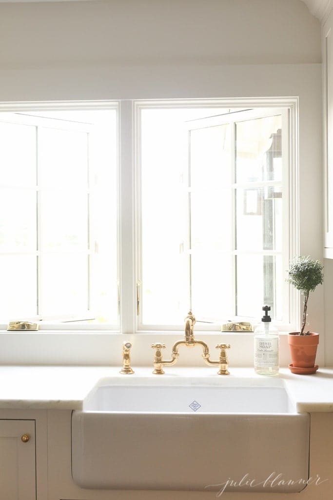 Farmhouse style sink underneath a big window with gold accents. 