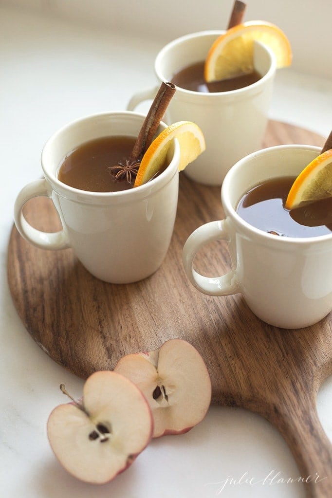 Spiked apple cider in white mugs garnished with cinnamon sticks and oranges on a wooden tray on a white countertop. 