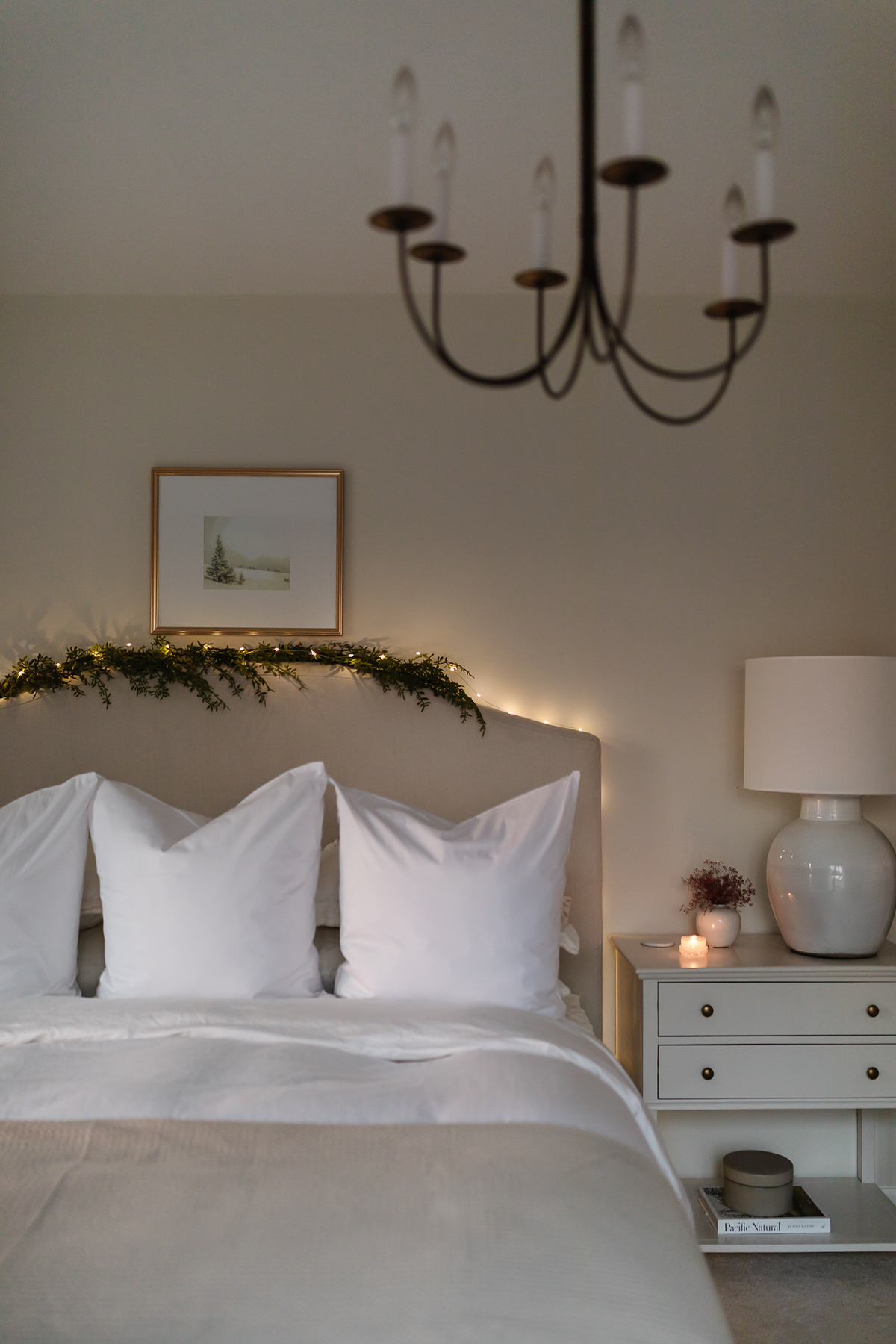 A bed adorned with white linens and a chandelier, illuminated by twinkling Christmas lights in the bedroom.