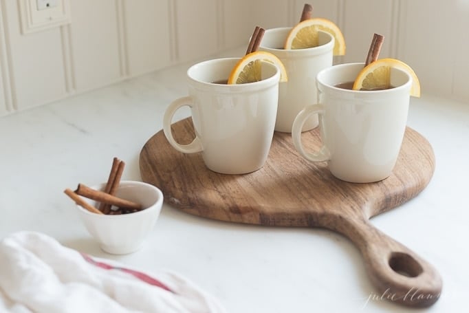 Spiked apple cider in white mugs garnished with oranges and cinnamon sticks on a wooden board. This is all on top of a white countertop. 