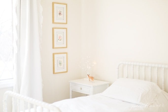 A white girl's bedroom with white blackout curtains and gold framed art.