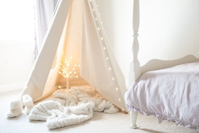A teepee in a white bedroom with a fairy light tree inside