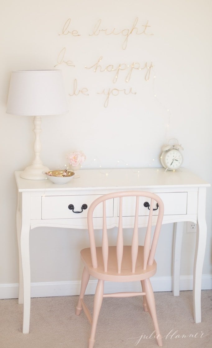 A girls' bedroom with a white lamp and gold wire words on the wall.