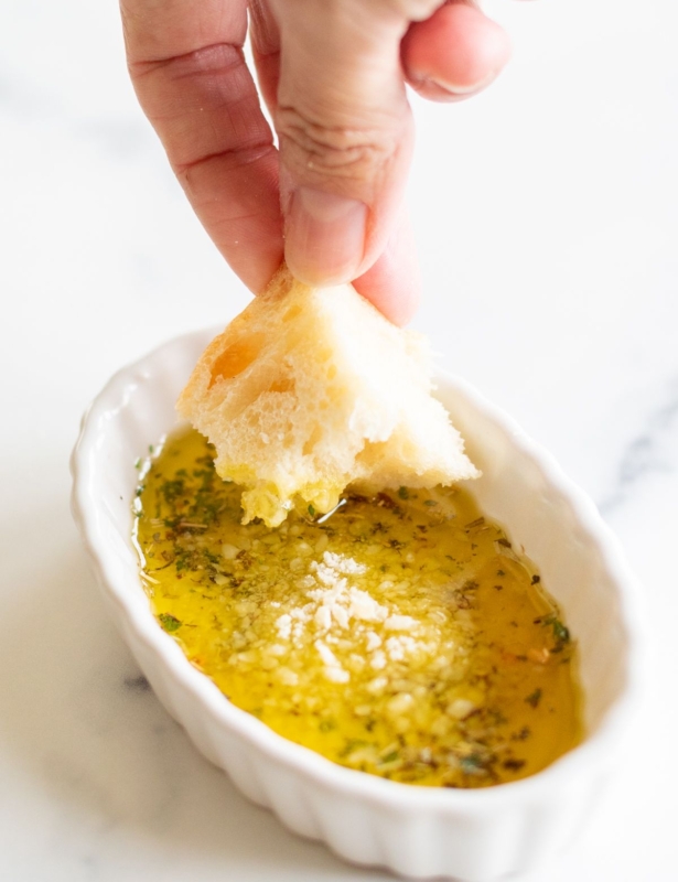 A hand dipping a piece of bread into a white ramekin full of olive oil bread dip.