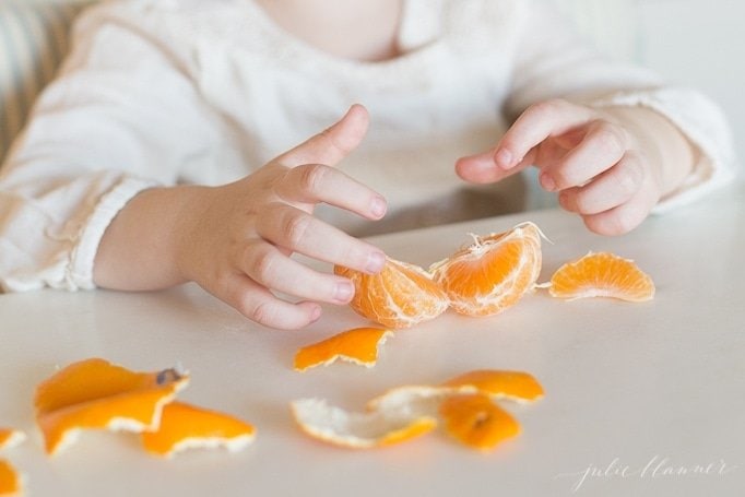 A child's hands peeling mandarin oranges on a white countertop.