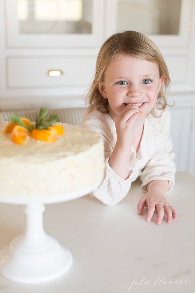 A little girl eating an orange with a homemade Mandarin Orange Cake Recipe in the foreground of image.