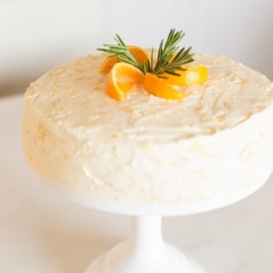 Sliced mandarin oranges and rosemary sprigs on top of a mandarin orange cake on a cake stand.