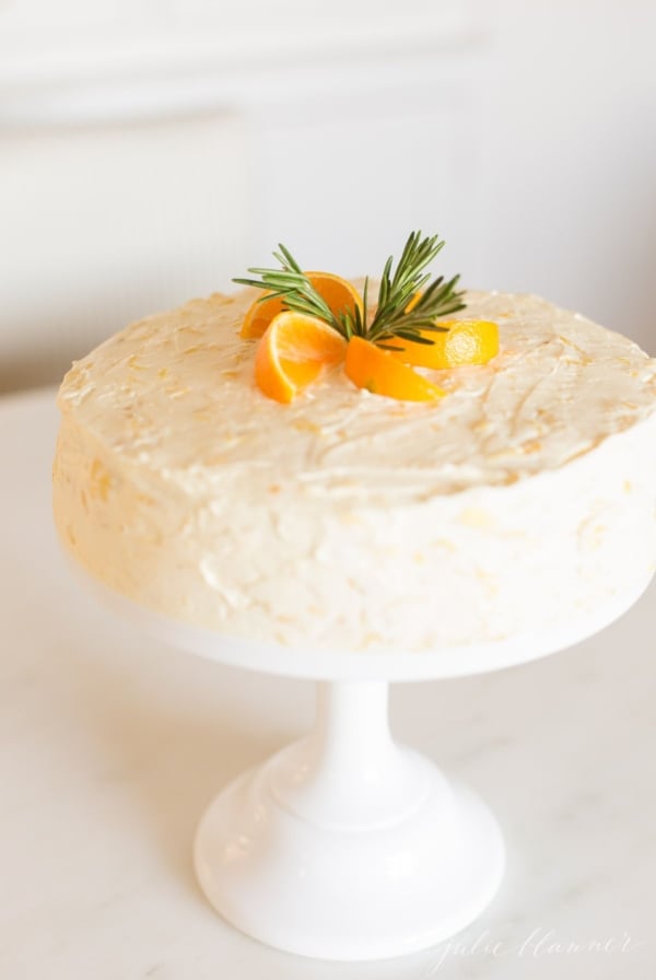 Sliced mandarin oranges and rosemary sprigs on top of a mandarin orange cake on a cake stand.
