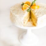 A round mandarin orange cake with a slice removed, placed on a white cake pedestal.