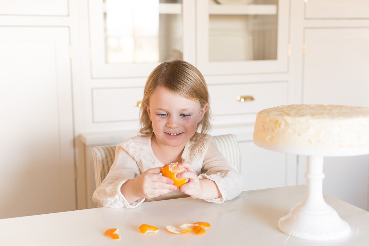 A little girl peeling mandarin oranges with a cake on a cake stand nearby.