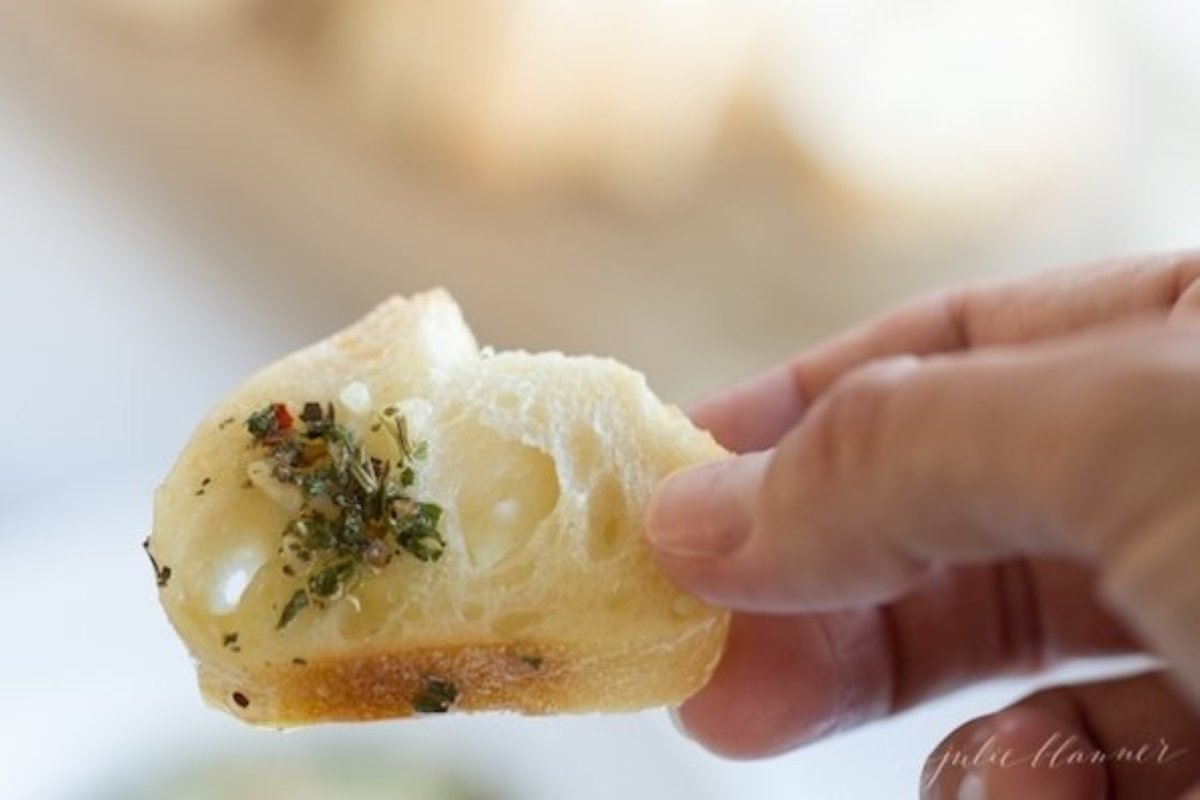 A hand holding a piece of crostini with olive oil and bread dipping spices.
