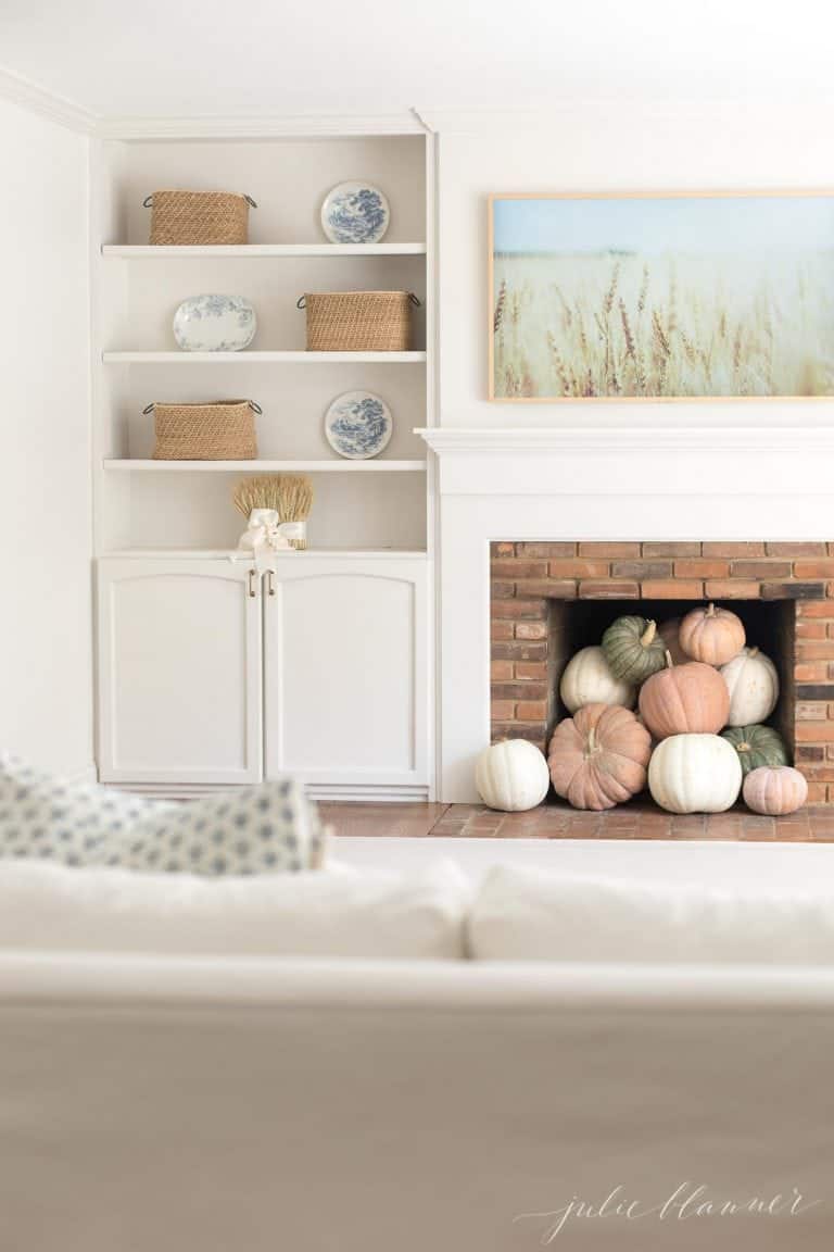 A white living room with a Samsung Frame TV that looks like art over the mantel, pumpkins inside fireplace and a fall image on the tv.