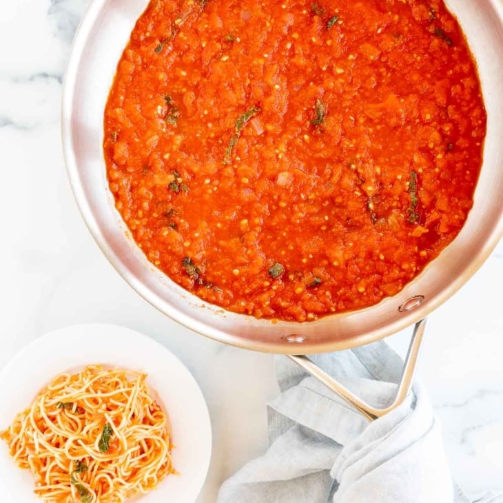 A silver saucepan full of pomodoro sauce, a plate of pomodoro pasta to the side