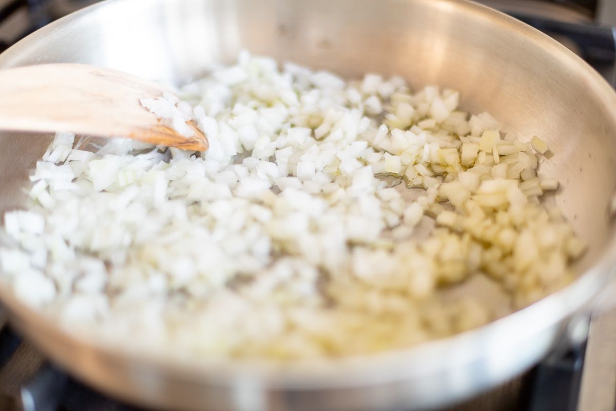 Chopped onions cooking in a silver pan with a wooden spoon.