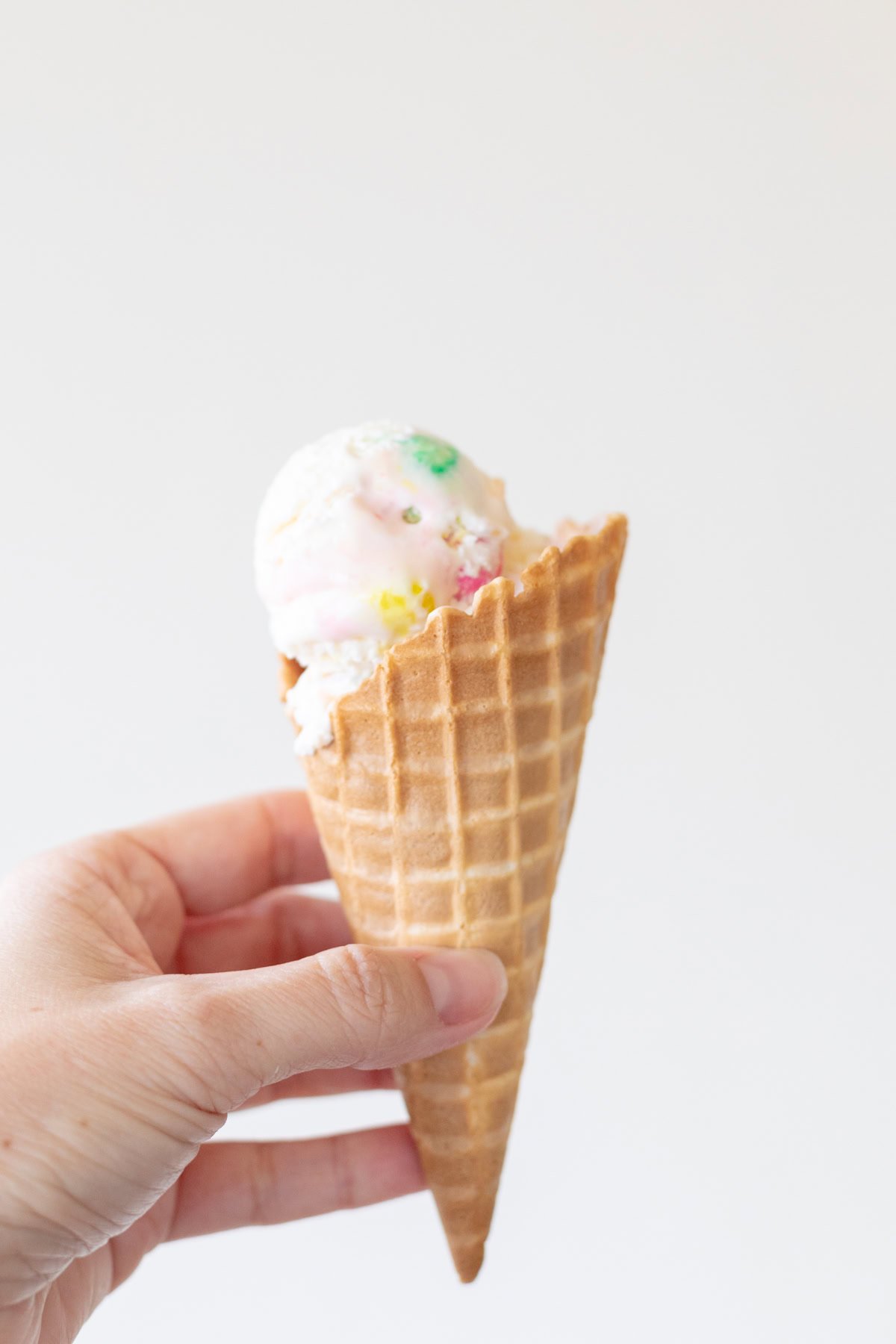 A hand holding a waffle cone with a single scoop of colorful bubble gum ice cream against a plain white background.