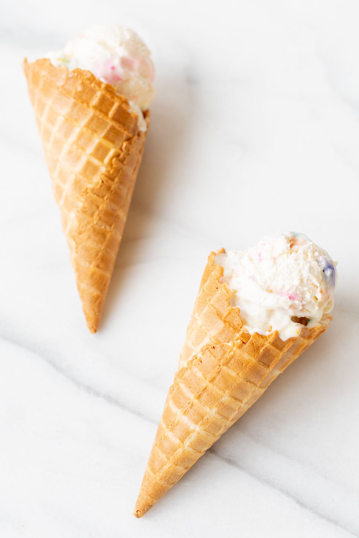 Two waffle ice cream cones with scoops of colorful bubble gum ice cream, placed on a marble surface.