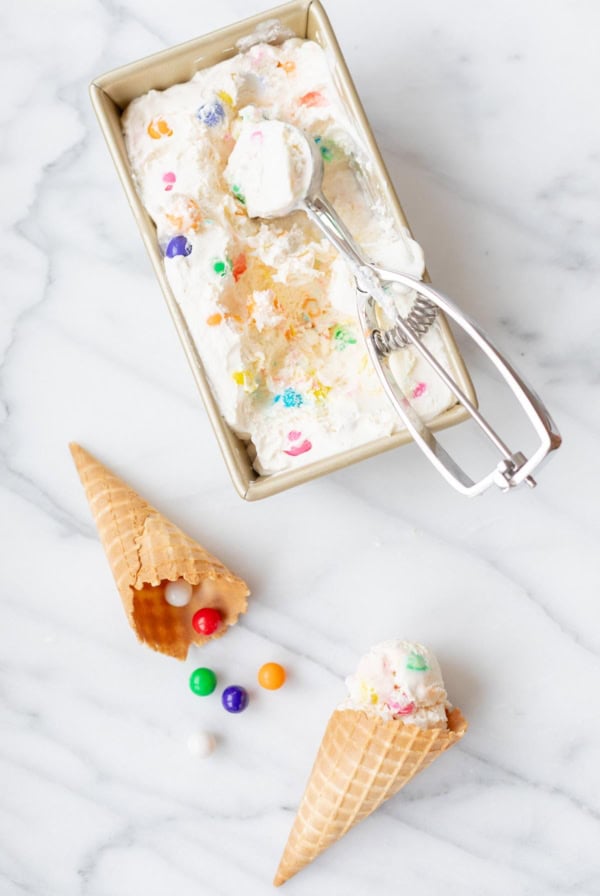 A rectangular container of bubble gum ice cream with colorful candy pieces, an ice cream scooper, two waffle cones—one boasting a scoop of ice cream and one overturned with candies spilling out—on a marble surface.