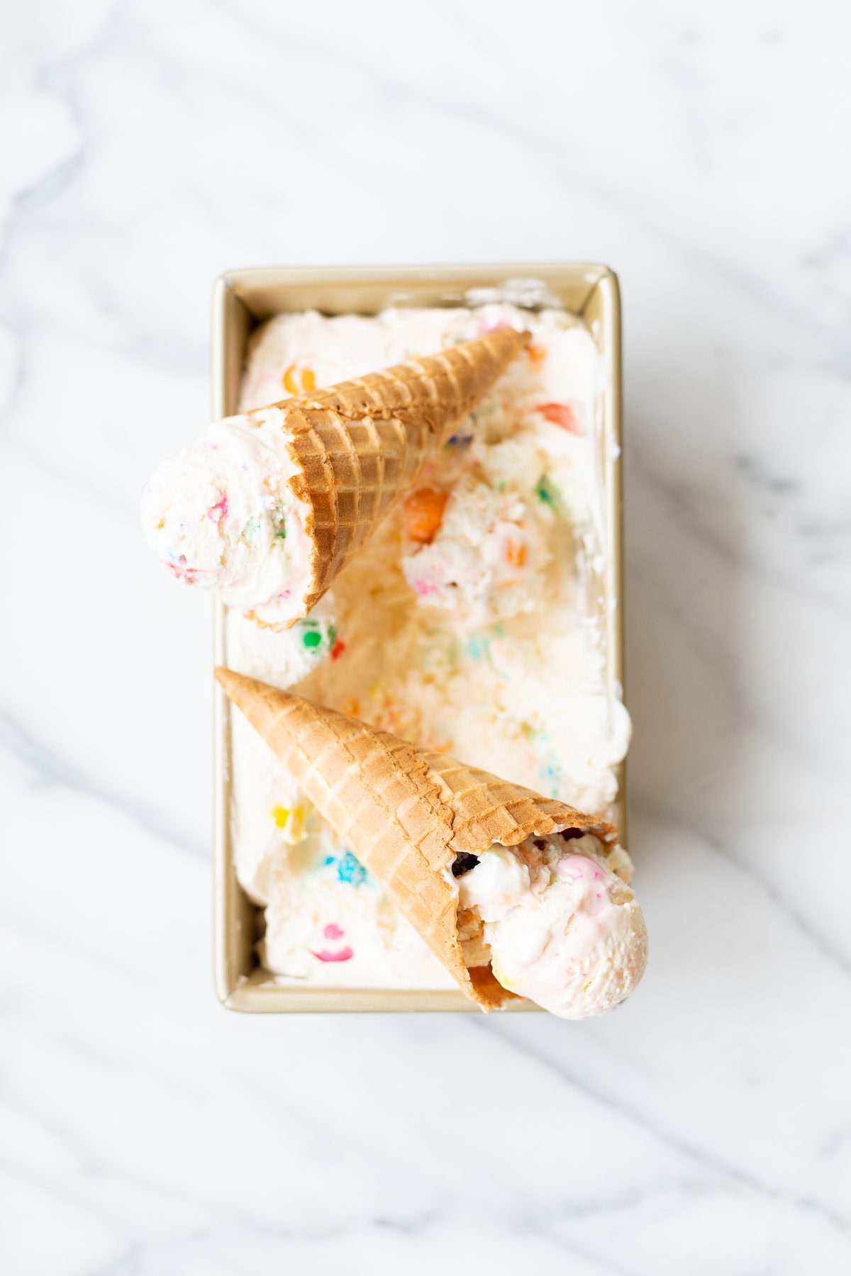 Two waffle cones filled with colorful, sprinkled bubble gum ice cream are placed on top of a rectangular container filled with the same ice cream, sitting on a white marble surface.