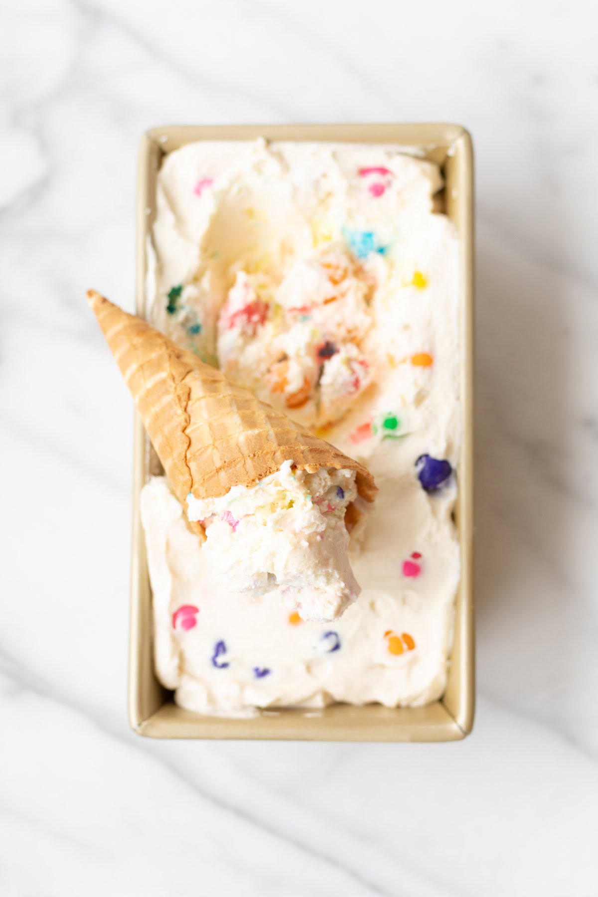 Rectangular tray of bubble gum ice cream with colorful sprinkles and an inverted ice cream cone placed on top, resting on a marble surface.
