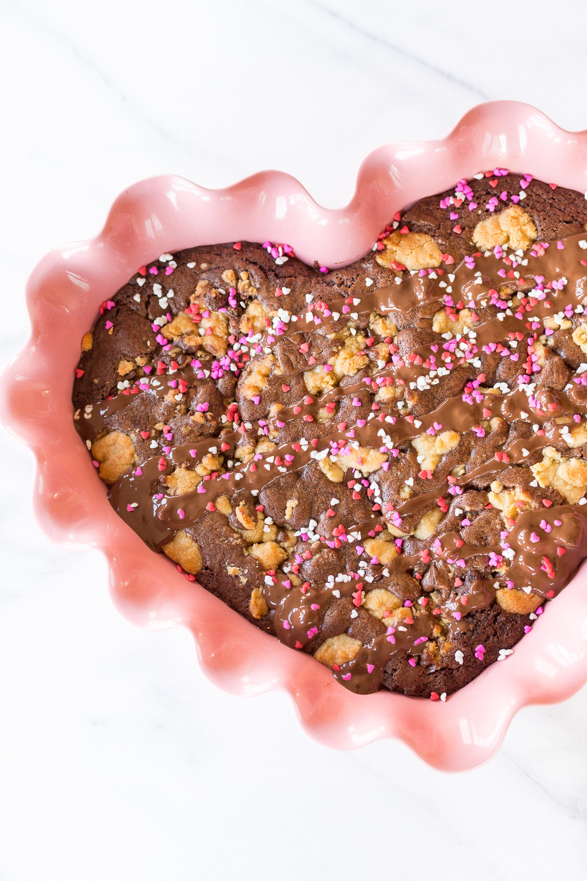 A heart-shaped chocolate brookie in a pink ceramic dish, topped with nuts and colorful sprinkles on a marble surface.