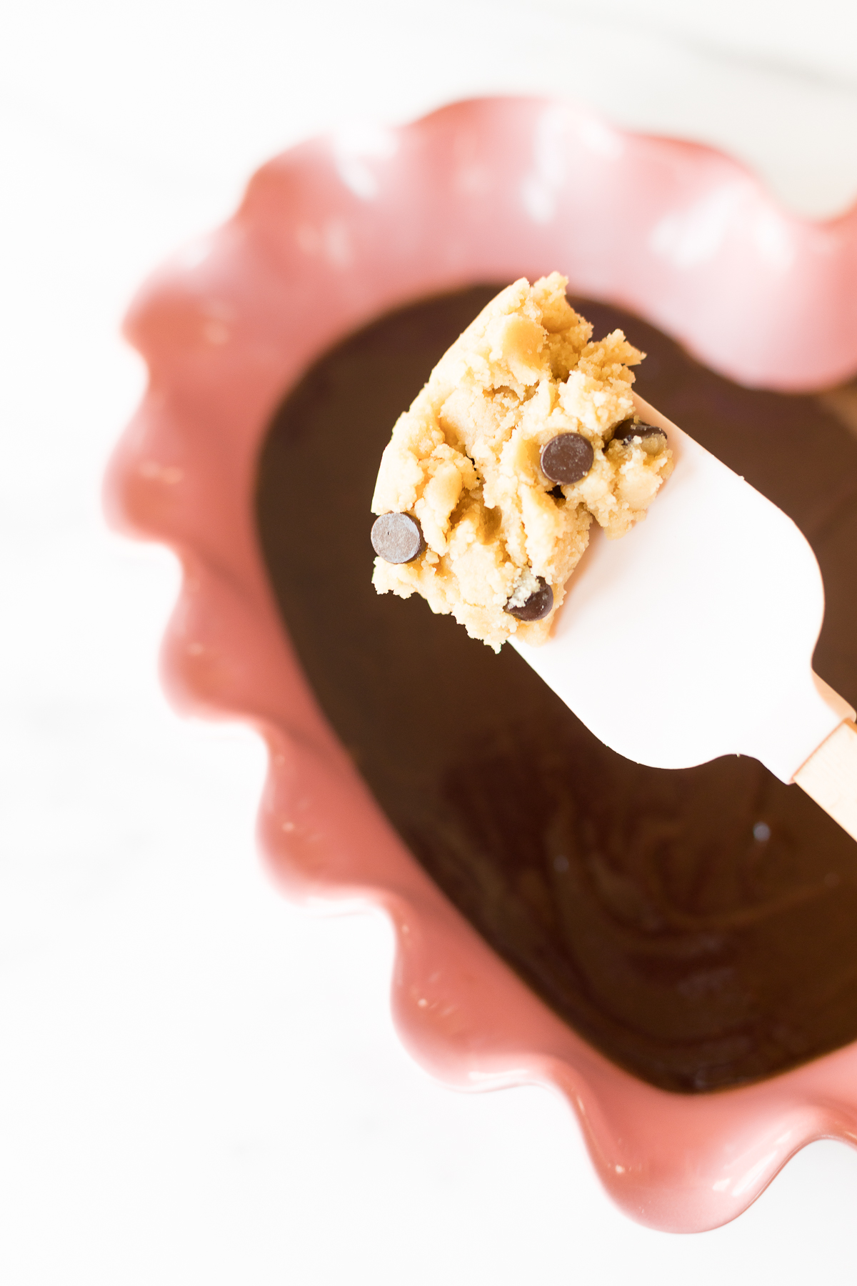 A scoop of brookie dough with chocolate chips on a white spatula, partially dipped in a bowl filled with melted chocolate.